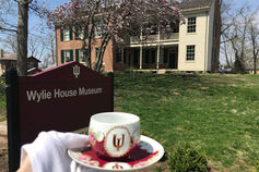 A crimson and white China teacup and saucer with pink flowers. The IU teacup is held up by a gloved hand next to an exterior building sign that reads, “Wylie House Museum.” A two-story brick home with a large porch is in the background.